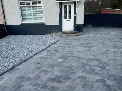The image shows a newly paved driveway made of grey interlocking pavers leading to the entrance of a house. To the left of the driveway is an area covered with grey gravel.