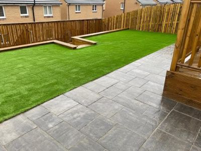 This image shows a backyard with artificial turf, stone paving, and a wooden fence. The backyard is neatly landscaped with a modern design.