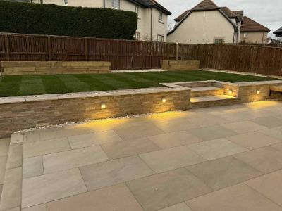 A newly landscaped backyard features a patio with stone walls, built-in lighting, and a tiered garden area with a fresh lawn. The garden is enclosed by wooden fences and landscaping features.
