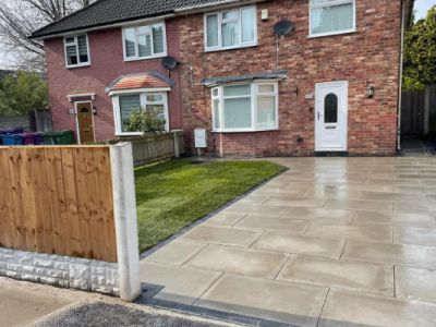 A newly paved driveway with stone slabs and a small patch of lawn is in front of a brick house. A wooden fence with stone base is on the left side of the image.