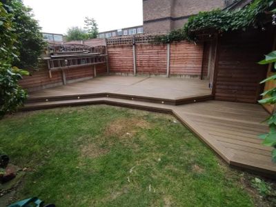 A newly built wooden deck with steps and integrated lighting is shown in a backyard garden. The deck extends from a small wooden shed and is surrounded by a wooden fence and lush green grass.