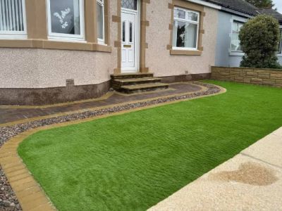 A newly landscaped garden features artificial turf with a paved pathway. A stone wall borders the garden area in front of a house.