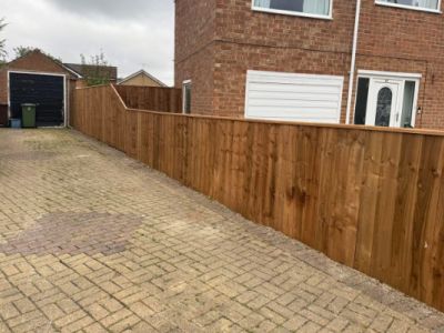 A wooden fence runs along a paved driveway in front of a brick house and garage.