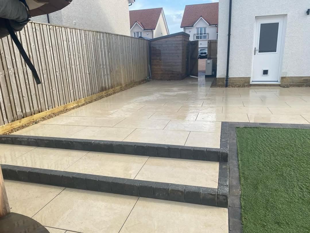 A newly paved patio with steps leads to a patch of artificial grass. A wooden fence borders the patio on one side, and a white house with a door is on the other.