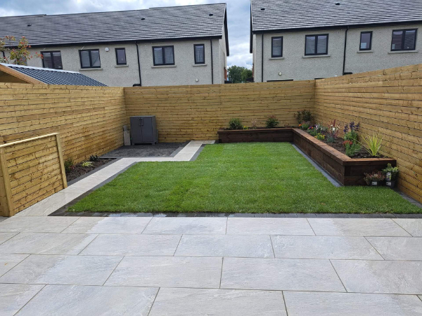 A newly landscaped backyard features a paved patio, a lush green lawn, and raised garden beds with plants. The area is enclosed by a modern wooden fence, with contemporary houses visible in the background.