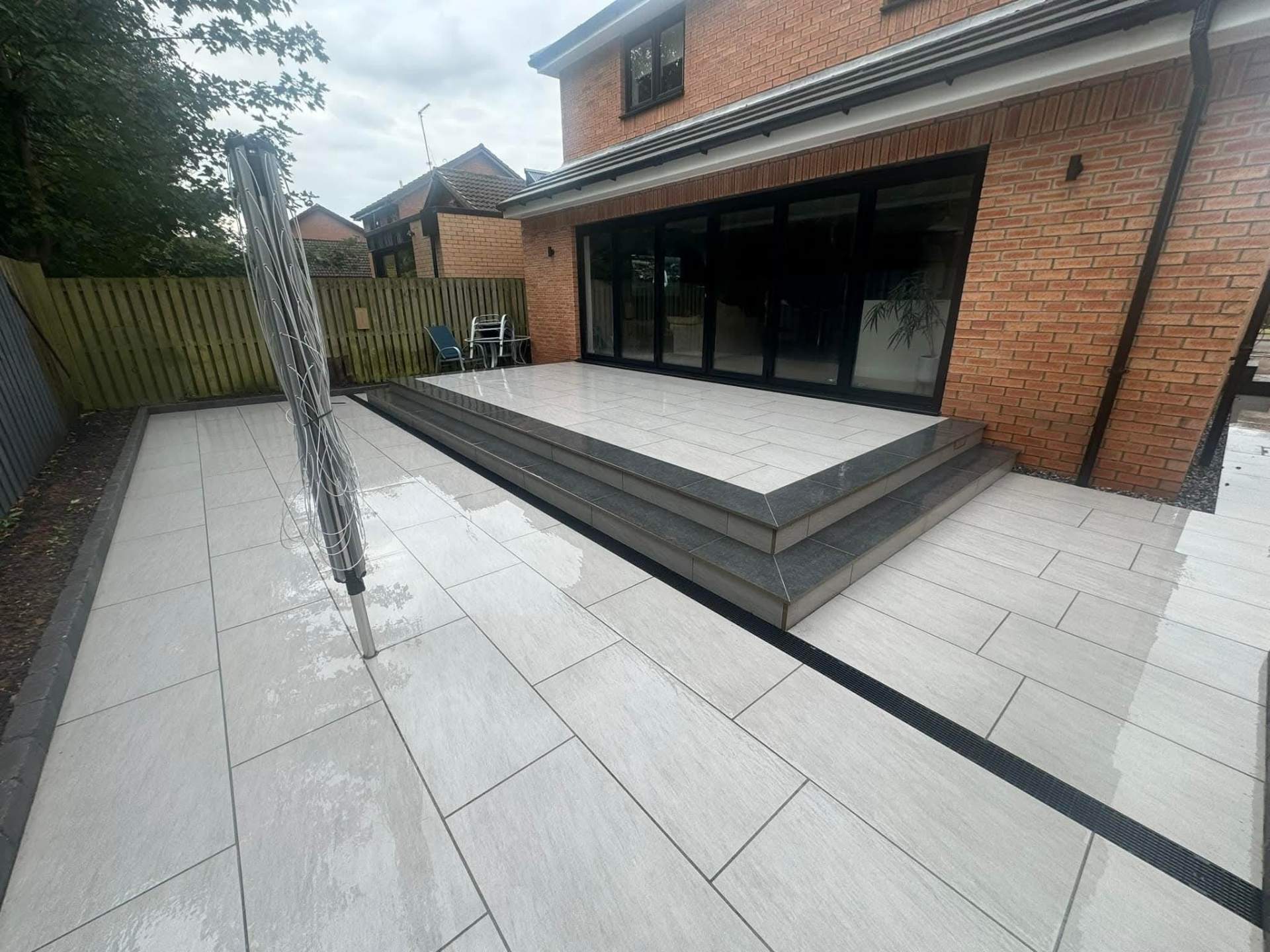 A modern patio area with light gray paving stones and a raised deck features bi-fold doors opening to a brick house. A rotary clothes line stands on the patio.