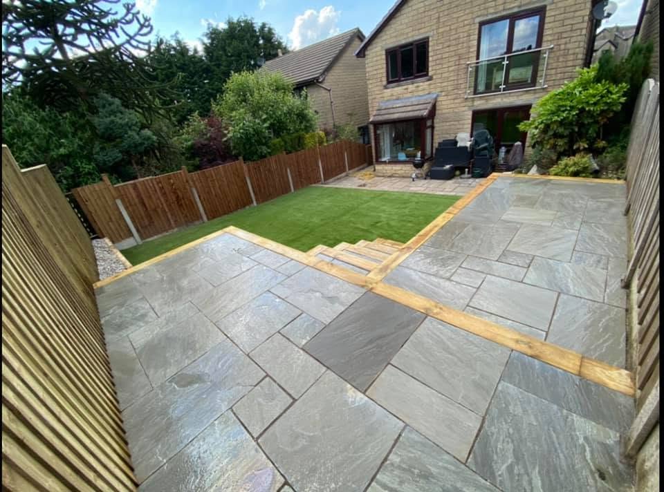 A newly paved patio with grey stone slabs leads to a small lawn area. Steps with wooden edging connect the patio to the lawn, all surrounded by a wooden fence.