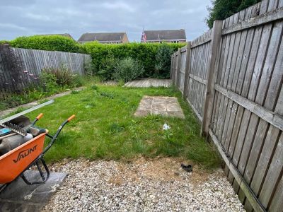 An overgrown garden with gardening tools in a wheelbarrow is pictured, with a wooden fence and houses in the background. The garden appears to be in a state of disarray with patches of weeds and grass.