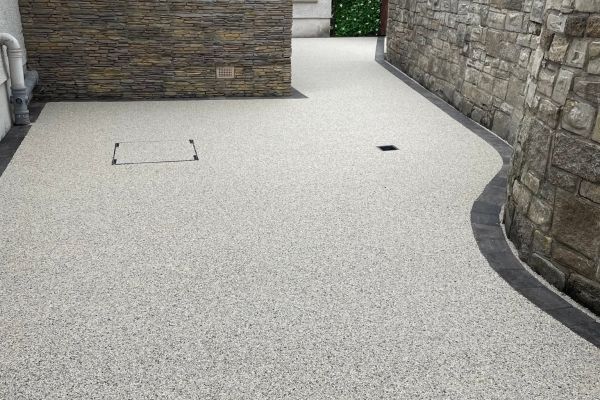 A textured resin driveway leads to a house with stone and modern cladding. The driveway is bordered by a stone wall and features a drainage grate.