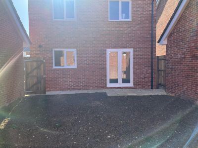 A red brick house with several windows and double doors is shown from the backyard. The backyard is mostly covered in dark soil and a small patio area.