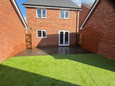 A red brick house with a patio and artificial turf lawn is shown in a backyard. The house has two stories with several windows and double doors leading to the patio.
