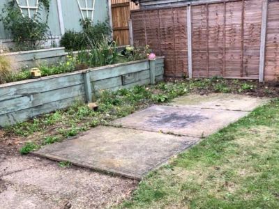 A neglected garden features a tiered wooden planter box with overgrown plants and weeds. Adjacent to the planter, there are concrete slabs and patchy grass, with a wooden fence in the background.