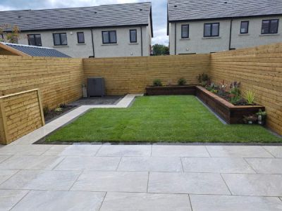 A newly landscaped garden features a modern paved patio, a lush green lawn, and raised wooden planters with various plants. The garden is enclosed by a horizontal wooden fence, with townhouses visible in the background.